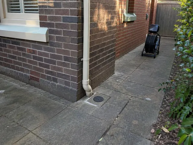 Capped drain inspection point beside a brick house side path with a drain camera reel and paved yard in Pymble