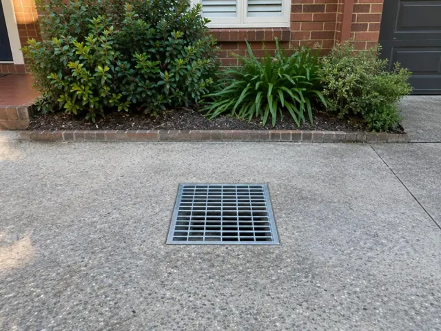 A flush stormwater grate set in a driveway beside brick garden edging and shrubs in Gordon