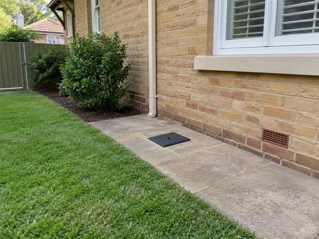 Flush black drain access cover in a paved side yard beside a brick house in Pymble